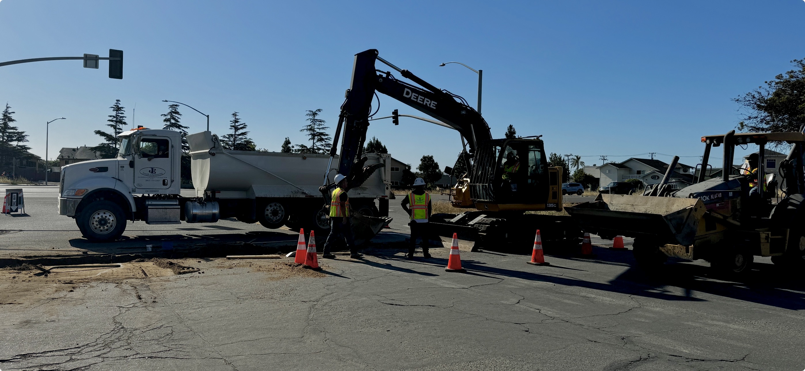 Joseph Engineering street utility work with dump truck, excavator on trailer, and crew in safety vests managing traffic on urban roadway